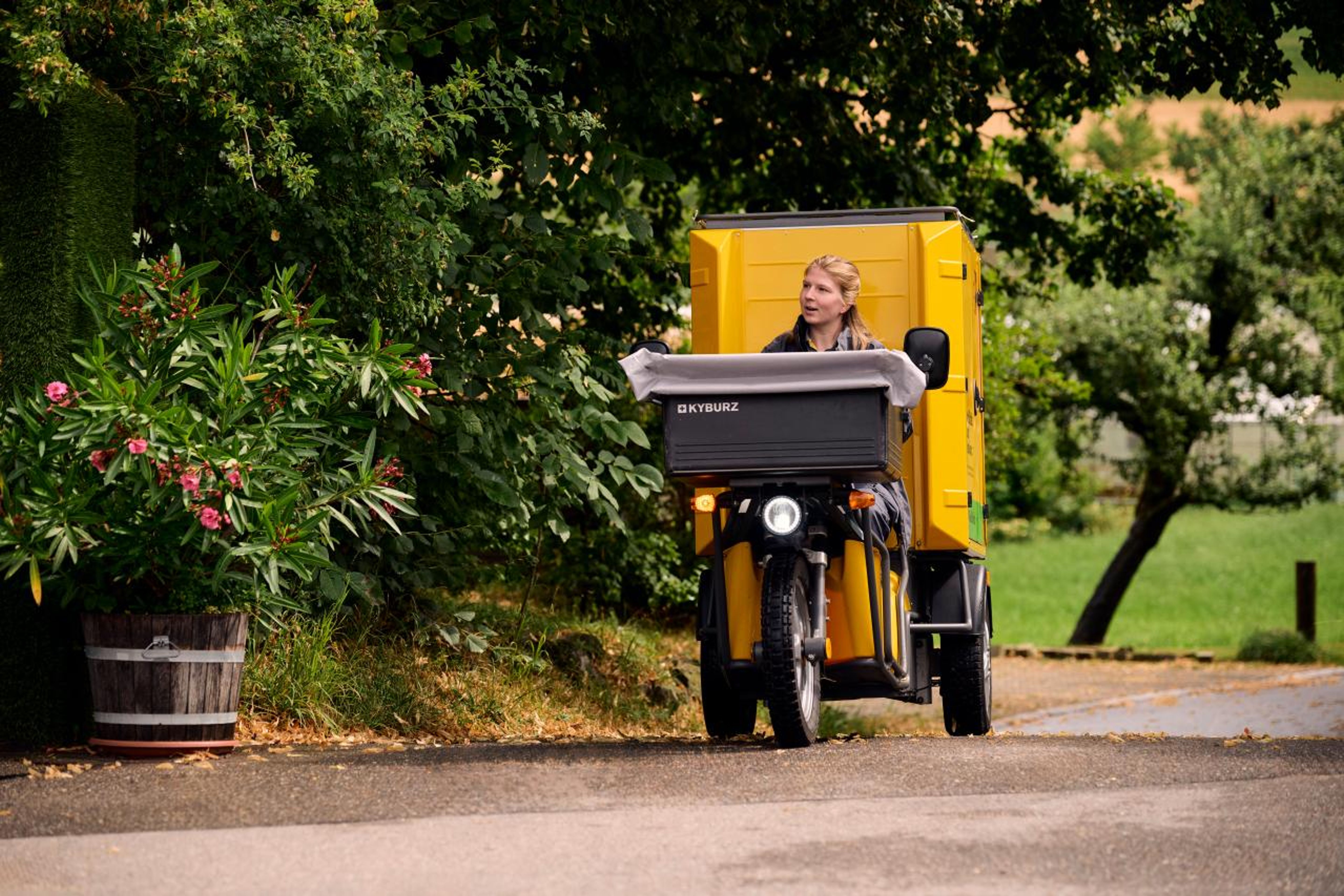 Eine Mitarbeiterin der Schweizerischen Post fährt auf einem dreirädrigen Elektromobil der Marke Kyburz entlang einer ländlichen Strasse. Das Fahrzeug ist gelb und mit einem Postlieferraum ausgestattet. Die Umgebung ist grün und malerisch, mit Bäumen und Blumentöpfen. Die Atmosphäre ist ruhig und idyllisch.,