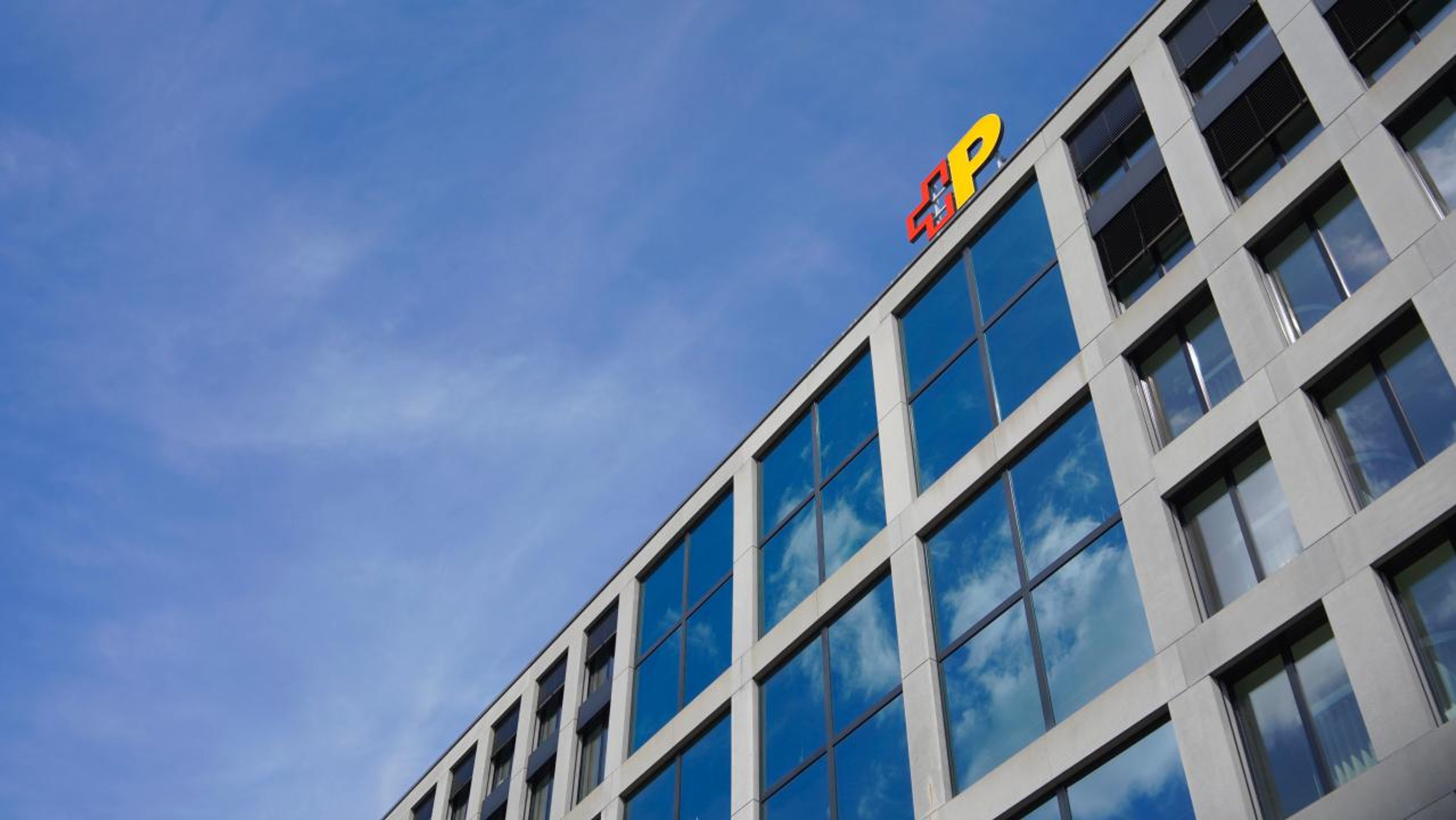Das Bild zeigt die Fassade eines modernen Bürogebäudes mit grossen Glasfenstern und einem Logo der Schweizerischen Post auf dem Dach. Der Himmel ist klar und blau, was die Architektur des Gebäudes unterstreicht.,