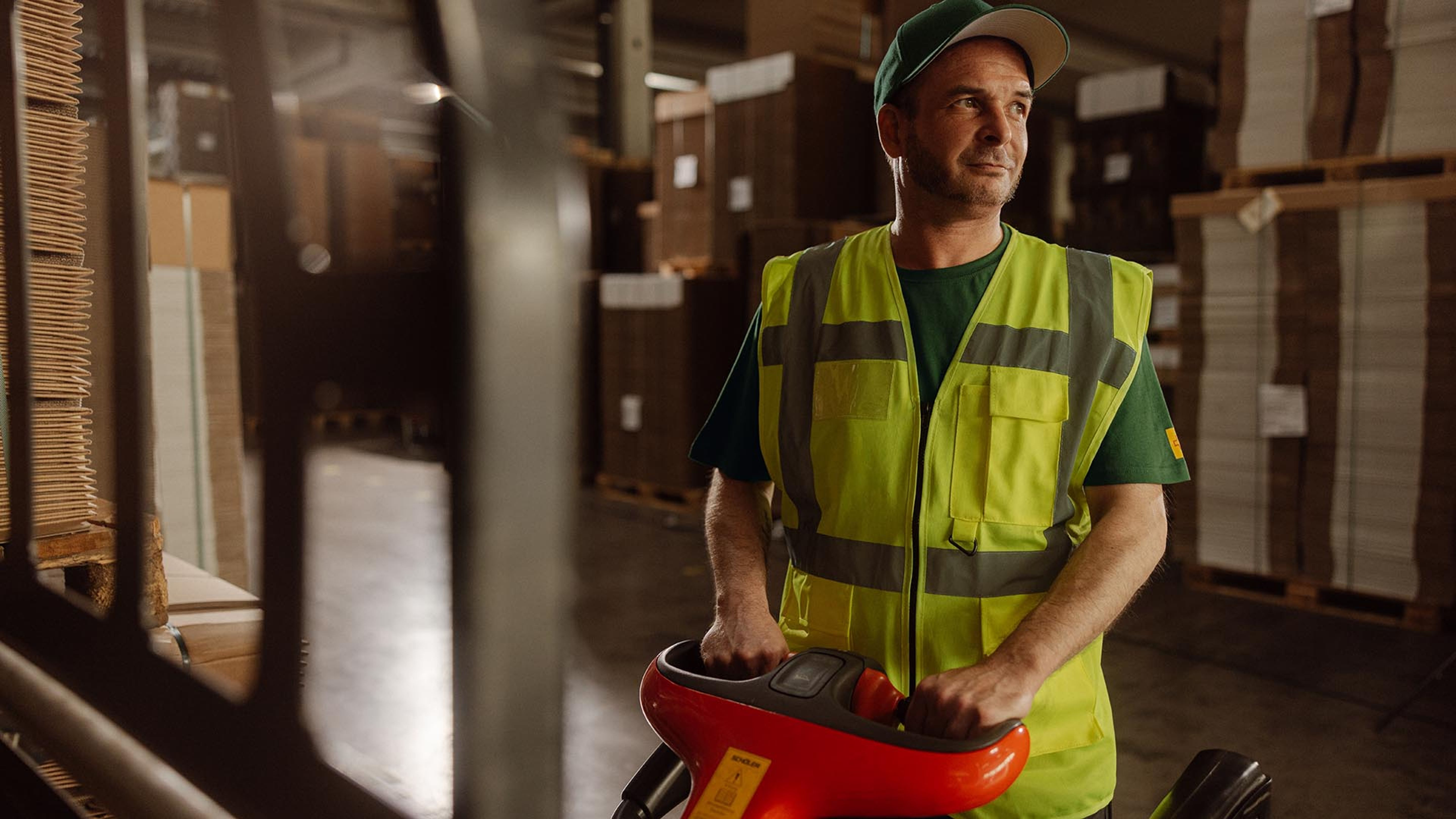 Un collaborateur vêtu d’une veste de sécurité utilise un chariot élévateur à plate-forme électrique dans un entrepôt et transporte des palettes de marchandises entre les rayonnages.